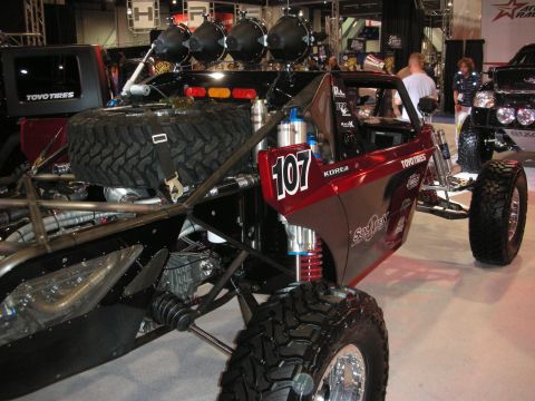 1998 Ford Crown Victoria - red and black tube-chassis off-road race buggy numbered 107 sits on display under bright lights at an indoor automotive show.