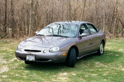 1996 Ford Taurus - A gray 1996 Ford Taurus sedan is parked on a grassy area with trees behind it.