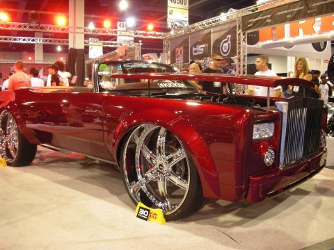 1998 Ford Crown Victoria - deep-red customized Rolls-Royce convertible with oversized chrome spinner wheels is displayed on a show stand surrounded by spectators.