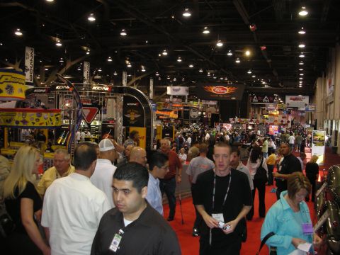 1998 Ford Crown Victoria - crowded indoor automotive trade show floor is filled with attendees walking among lit vendor booths and overhead banners.