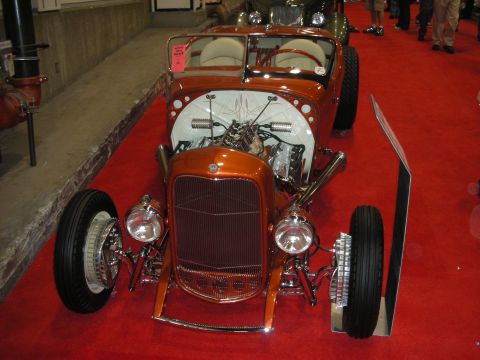 1998 Ford Crown Victoria - orange hot rod roadster with an exposed chrome engine, chrome grille and wire wheels sits on red show carpet.