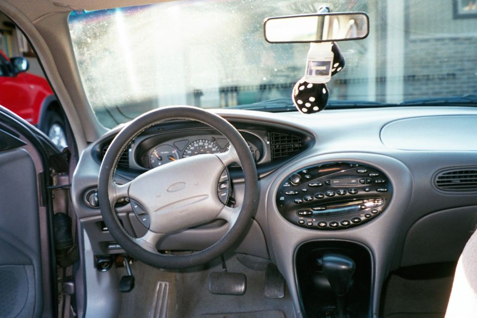 Gray Ford Taurus interior with Ford steering wheel, oval center stack radio, and fuzzy dice hanging from mirror.