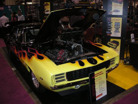 1998 Ford Crown Victoria - black first-generation Camaro with yellow flame paint sits with its hood up showing a tall blown engine at an indoor show.