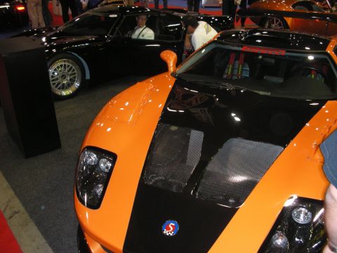 1998 Ford Crown Victoria - orange and black sports car with a meshed carbon hood and a small badge sits beside a black supercar at an indoor show.