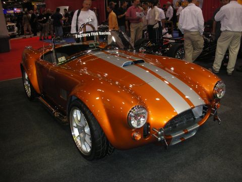 1998 Ford Crown Victoria - copper-orange Cobra-style roadster with silver racing stripes and polished wheels sits at a Factory Five Racing booth.