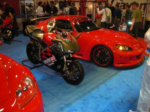 1998 Ford Crown Victoria - red roadster parked beside a black and silver Yamaha sportbike on blue show carpet, with vendor booths behind.