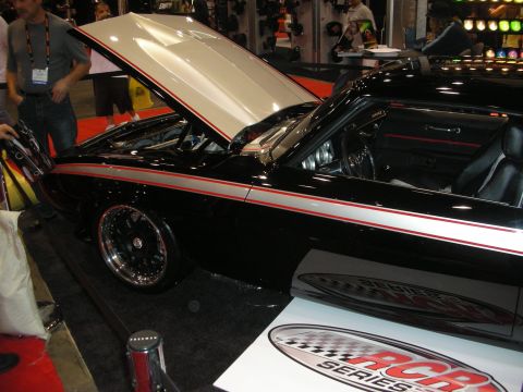 1998 Ford Crown Victoria - black muscle car with a red pinstripe and open hood sits on display near an RCR Series sign at an indoor show.