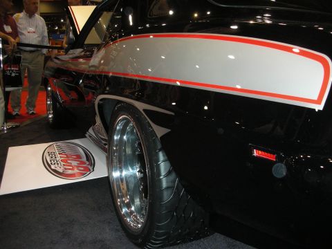 1998 Ford Crown Victoria - Close-up of a black custom muscle car's rear quarter panel with a red-bordered white stripe and polished five-spoke wheel at an indoor expo.
