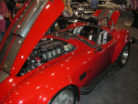 1998 Ford Crown Victoria - Red Cobra-style roadster displayed with its hood open exposing a chrome-trimmed V8, polished wheels and a chrome roll bar at an expo.