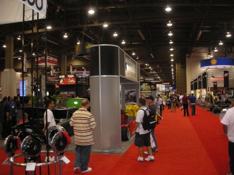 1998 Ford Crown Victoria - Wide view down a busy trade-show aisle with red carpet, exhibitor booths and a foreground display of brake rotors and calipers.