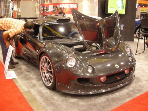 1998 Ford Crown Victoria - bare carbon-fiber Lotus-style roadster with its clamshell front bodywork raised, on display in a booth with onlookers.