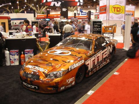 1998 Ford Crown Victoria - orange camouflage-wrapped Scion race car covered in sponsor decals including Titan and Fidanza, displayed on a show floor.