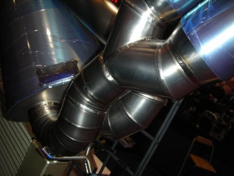 1998 Ford Crown Victoria - Detail of a welded titanium exhaust collector joining ribbed pipes to two blue-tinted mufflers on a display stand.