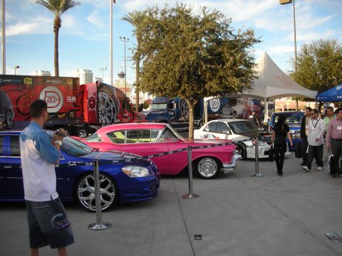 1998 Ford Crown Victoria - Outdoor car-show lot with a blue customized car, a pink vintage 1950s coupe, and spectators near a tent.