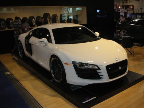 1998 Ford Crown Victoria - White Audi R8 mid-engine sports car displayed on a platform at an indoor auto show with spectators behind.