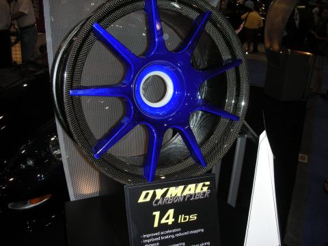 1998 Ford Crown Victoria - Carbon-fiber wheel with blue spokes displayed beside a Dymag sign reading '14 lbs' at a trade show.