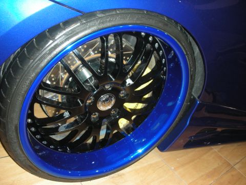 1998 Ford Crown Victoria - Close-up of a black multi-spoke wheel with a blue outer lip mounted on a blue car over a tile floor.