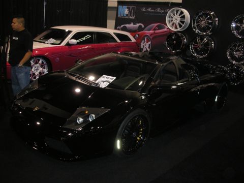1998 Ford Crown Victoria - Black Lamborghini Murcielago parked before a wall of chrome wheels and a red car at an indoor show booth.