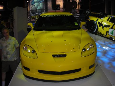 1998 Ford Crown Victoria - bright yellow Lingenfelter-badged Corvette sits on display at an indoor auto show with a yellow C6.R race car behind it.