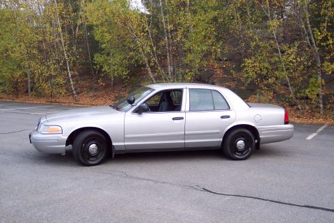 2001 Ford Crown Victoria - Side view of a silver 2001 Ford Crown Victoria sedan with black wheels and a police light on the driver's side mirror.