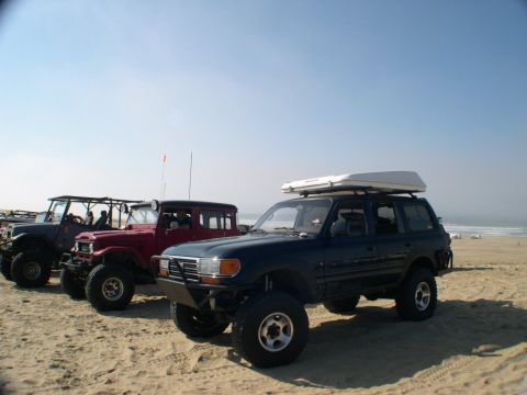 2007 Toyota FJ Cruiser - Several off-road vehicles, including a dark blue SUV with a rooftop tent, are parked on a sandy beach under a clear blue sky.