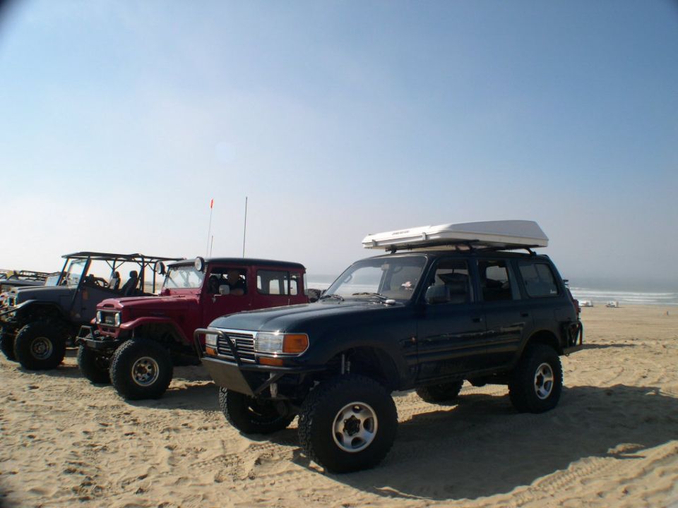 Lifted dark blue Toyota 80-series Land Cruiser with roof box on a beach beside two FJ40s