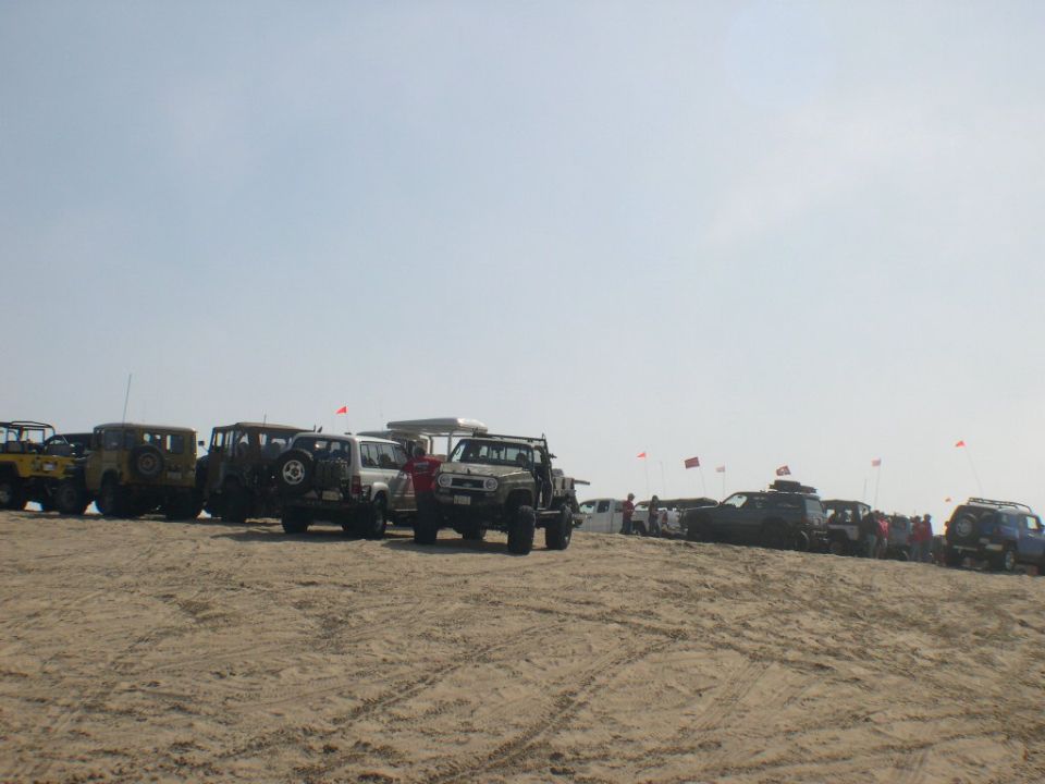 Row of vintage Land Cruisers and 4x4s with red whip flags parked on a sand dune