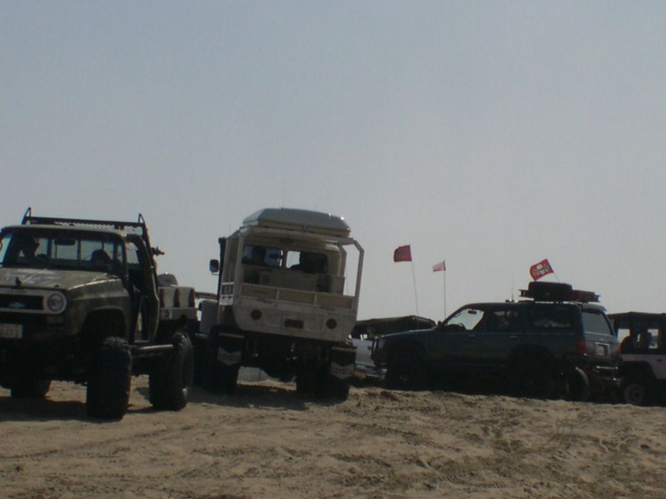 Olive 4x4, tan military-style flatbed truck and blue Land Cruiser parked on sand dunes
