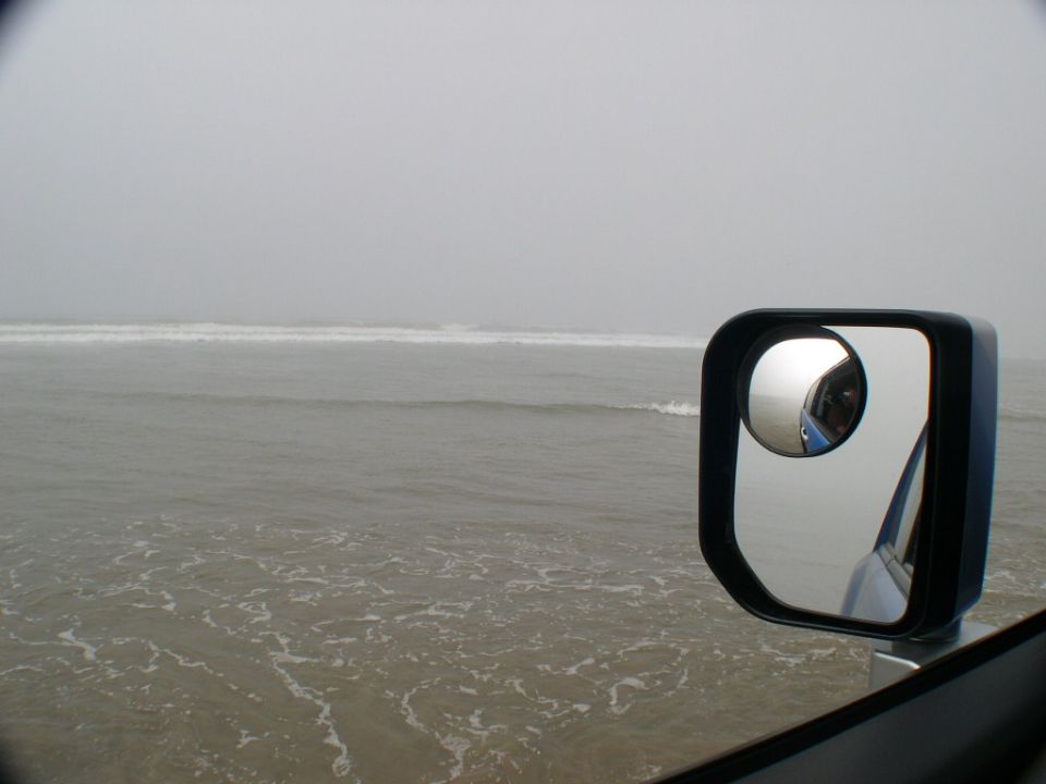 Square black side mirror with convex blind-spot lens on a silver 4x4 beside foggy surf