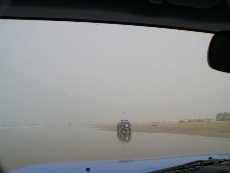 View over a blue hood of a dark 4x4 with whip flag driving on wet foggy beach sand