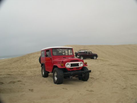 2007 Toyota FJ Cruiser - Two Toyota FJ Cruisers, one classic and one modern, are parked on sand dunes near the ocean.