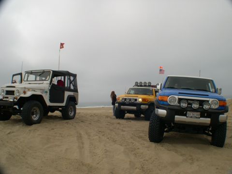 2007 Toyota FJ Cruiser - Three off-road vehicles parked on a beach under a cloudy sky, with flags flying in the background.