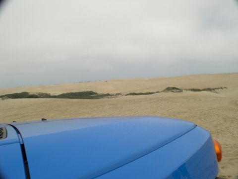 2007 Toyota FJ Cruiser - A blue SUV's hood and turn signal are visible in the foreground, with sand dunes stretching into the distance under an overcast sky.