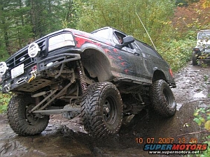 1986 Ford Bronco - A mud-caked lifted full-size Bronco flexes on a rocky, wet trail with the front driver-side wheel climbing as the suspension articulates. I…