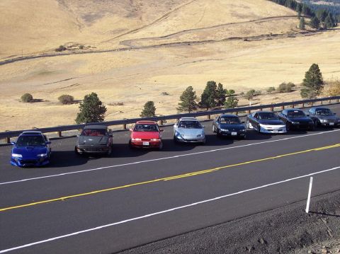 1995 Ford Probe - A collection of coupes are lined up along a scenic roadside, with a vast, arid landscape stretching out behind them.