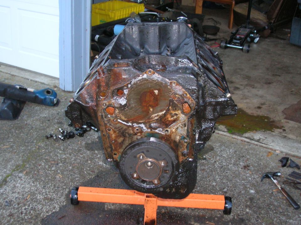 Engine block on an orange stand, with rust and oil visible. Tools and a jack are in the background
