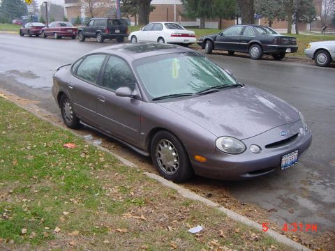 1996 Ford Taurus - A 1996 Ford Taurus is parked on a wet street, with other vehicles visible in the background.