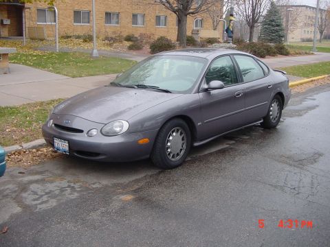 1996 Ford Taurus - A gray 1996 Ford Taurus sedan is parked on a wet street next to a sidewalk.
