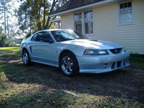 1999 Ford Mustang - A silver Ford Mustang with aftermarket chrome wheels is parked in front of a house.