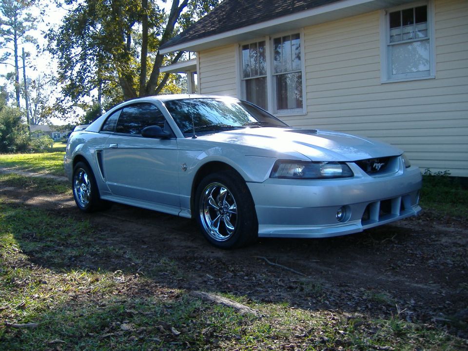 Silver Ford Mustang with chrome rims parked on a dirt and grass area in front of a light yellow house