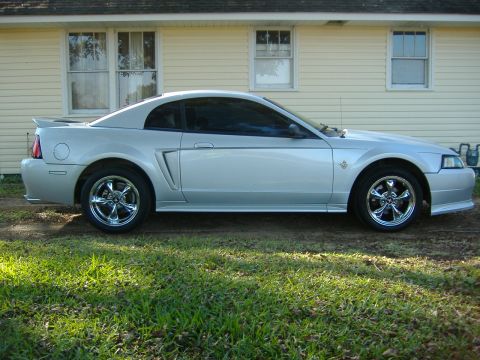 1999 Ford Mustang - A silver Ford Mustang is parked on a dirt patch in front of a yellow house.