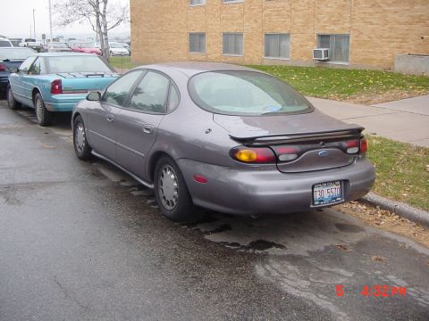 1996 Ford Taurus - A 1996 Ford Taurus is parked on a wet street next to another car, with a brick building in the background.