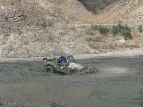1987 Suzuki Samurai - A white 1987 Suzuki Samurai navigates through a muddy terrain, with its tires deep in the mire.