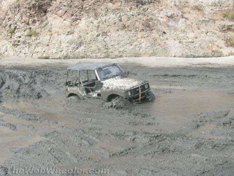 1987 Suzuki Samurai - A white 1987 Suzuki Samurai is deeply stuck in a muddy terrain, with only its upper body visible above the mire.