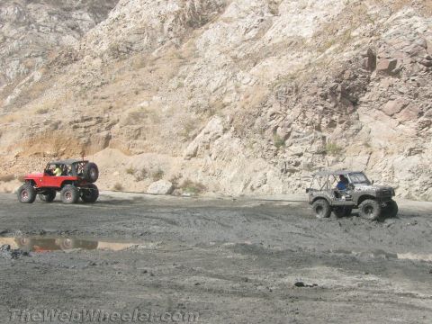 1987 Suzuki Samurai - Two off-road vehicles navigate a muddy terrain with a rocky hillside.