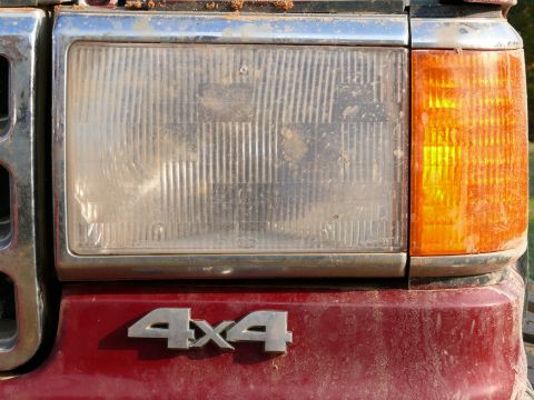 1988 Ford Bronco - Close-up of a red vehicle's front end, showing the headlight, turn signal, chrome grille, and a 4x4 badge.