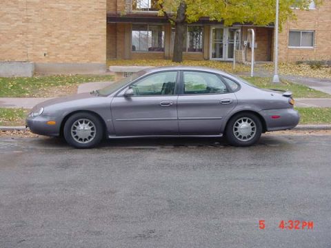 1996 Ford Taurus - A gray 1996 Ford Taurus sedan is parked on the side of a street next to a brick building.