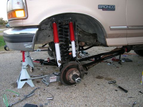 1996 Ford Bronco - Front suspension of a tan Ford Bronco XLT torn down with the wheel off, showing dual red-and-white shocks, a black coil spring, black radiu…
