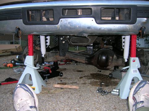 1996 Ford Bronco - Head-on view under the chrome front bumper of a truck raised on red jack stands, showing the Dana-style front axle housing, coil springs an…