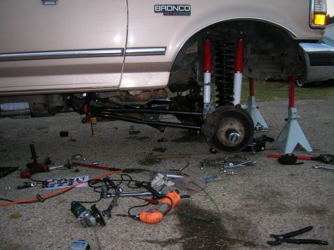 1996 Ford Bronco - Tan Ford Bronco XLT up on jack stands with the front wheel removed, exposing dual red-bodied shocks, black coil spring, black radius arm an…